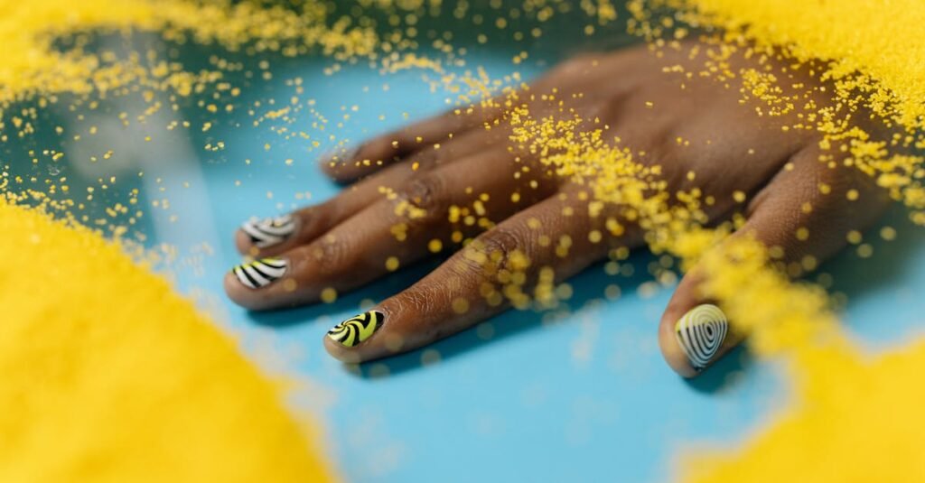 Artistic nail design with yellow and black patterns, surrounded by vibrant yellow sand on a blue surface.